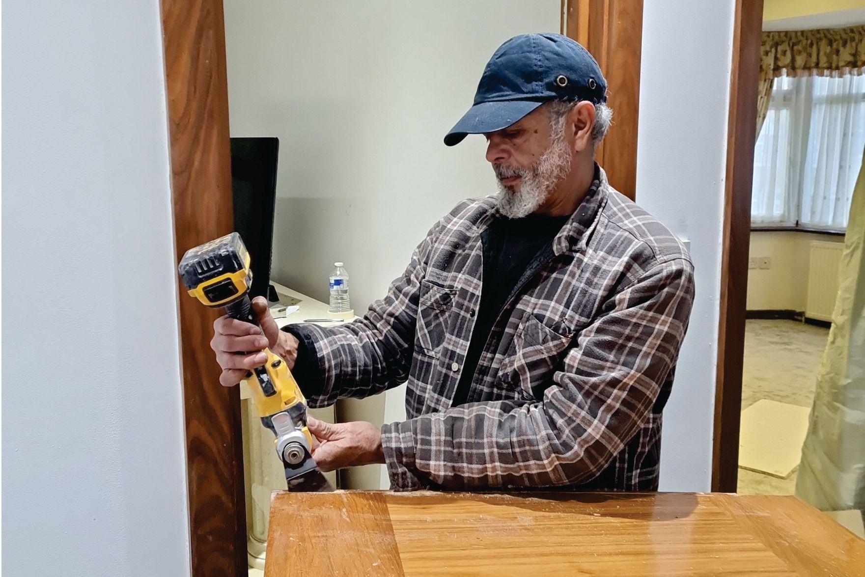 Person wearing a plaid shirt and cap using a power tool to sand a wooden surface indoors.