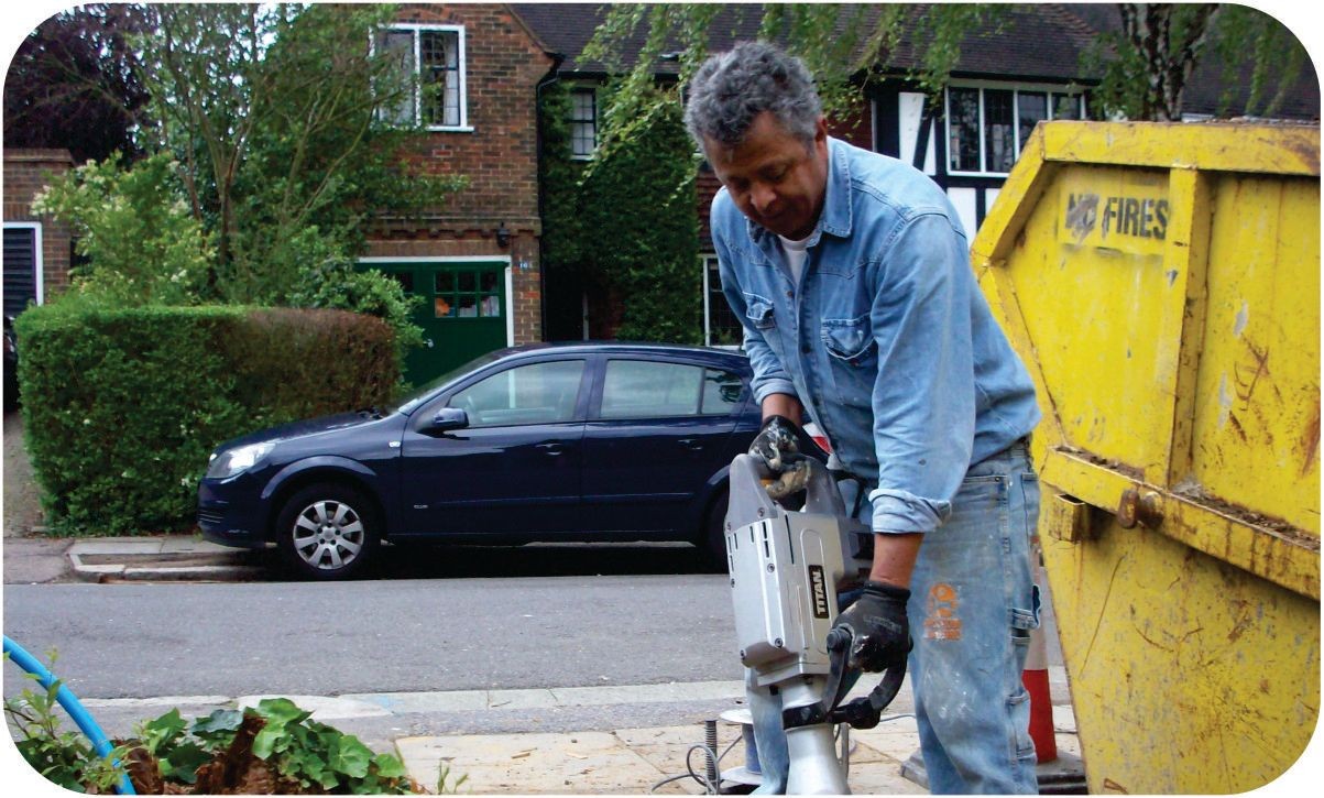 Man wearing a denim shirt using a power tool next to a yellow skip on a residential street.