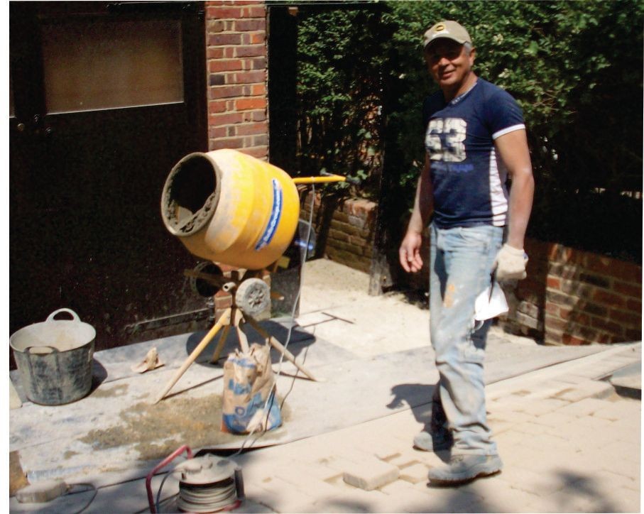 Man standing next to a yellow cement mixer on a construction site with various building materials around.